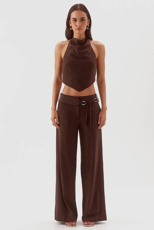 A model stands against a light gray studio background wearing a dark brown two-piece outfit consisting of a sleeveless halter-style crop top and matching wide-leg pants with a drawstring waist and metal ring detail.