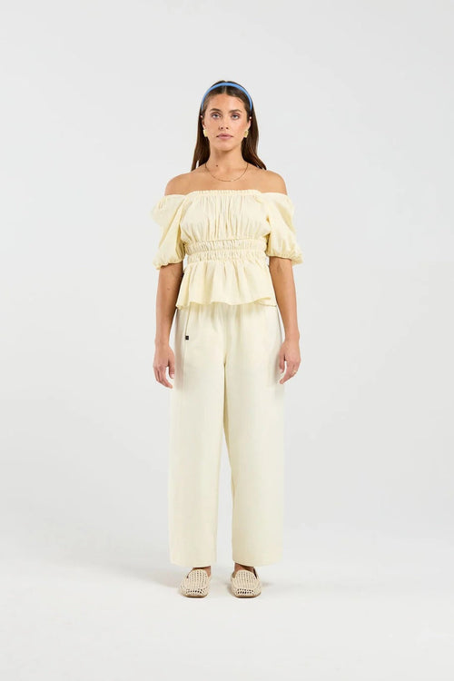 A woman modeling a pale yellow off-the-shoulder top with puff sleeves and matching wide-leg pants, standing against a plain light gray studio background.
