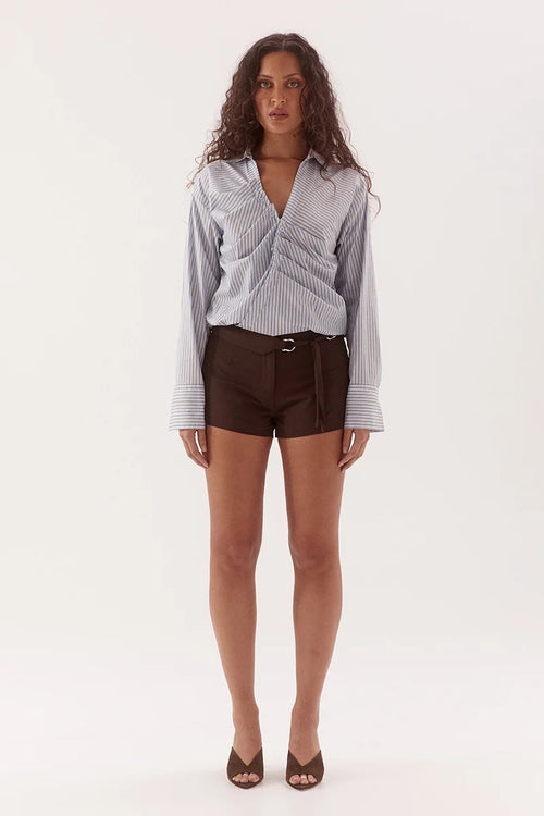 A woman stands against a plain white studio background wearing a light blue and white vertically striped long-sleeve wrap-style blouse tucked into dark brown shorts with a belt, paired with brown heeled sandals.