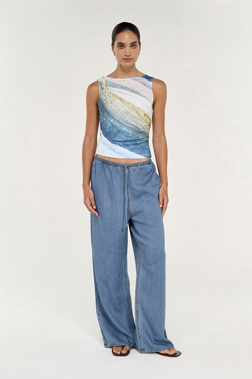 A model stands against a light gray studio background wearing a sleeveless cropped top with an abstract blue, beige, and white brushstroke pattern, paired with wide-leg blue jeans and dark sandals.
