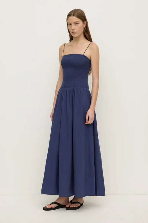 A woman modeling a sleeveless navy blue maxi dress with thin spaghetti straps, a smocked bodice, and a flowing A-line skirt, paired with black sandals against a plain off-white studio background.