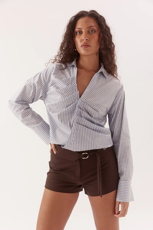 A woman modeling a light blue and white vertically striped wrap-style button-down shirt with dark brown shorts, photographed against a plain white studio background.