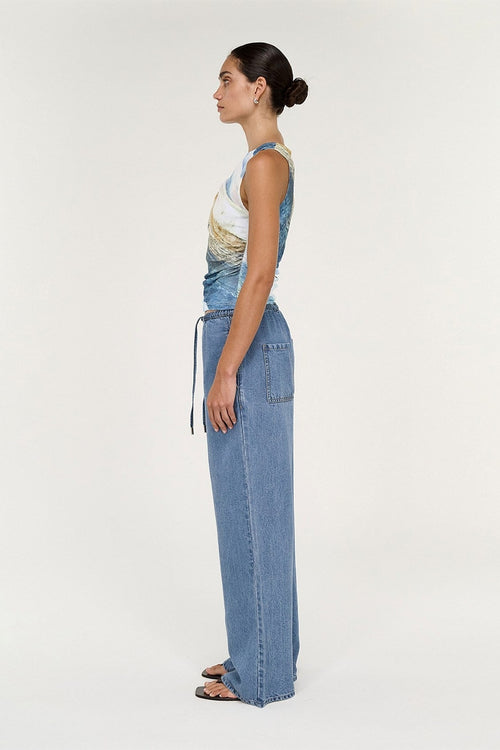A woman stands in profile wearing a sleeveless top with a blue, white, and beige landscape print and wide-leg blue denim pants, photographed against a plain light gray studio background.