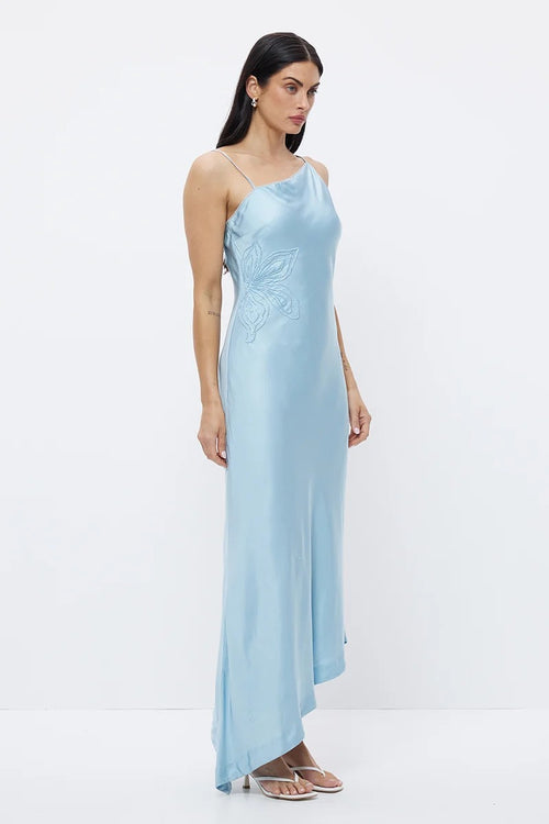A woman modeling a light blue satin one-shoulder maxi dress with delicate floral embroidery on the bodice, set against a plain white studio background.