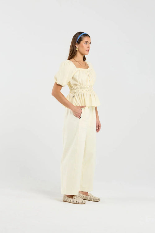 A woman modeling a cream-colored two-piece outfit consisting of a smocked peplum top with puff sleeves and matching wide-leg trousers, styled with a blue headband and woven flat shoes against a plain white studio background.