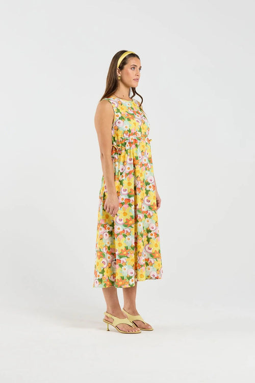 A woman stands in profile wearing a sleeveless midi dress with a bright multicolor floral print on a yellow base, paired with tan heeled sandals and a yellow headband, against a plain light gray studio background.