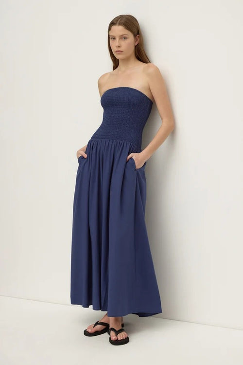 A woman modeling a strapless navy blue maxi dress with a smocked bodice and side pockets, standing against a plain off-white wall.