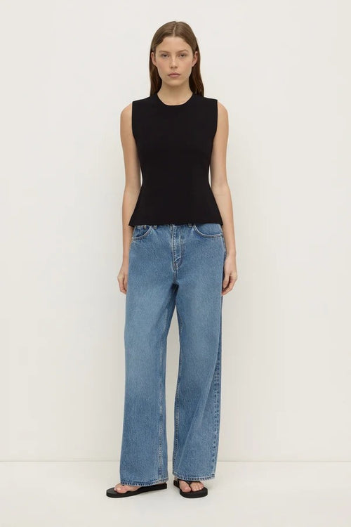 A model stands against a plain white studio background wearing a sleeveless black top, wide-leg light blue jeans, and black sandals.