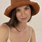 A woman wearing a brown woven sun hat with a wide brim, paired with a light beige spaghetti-strap top, photographed against a neutral beige background.