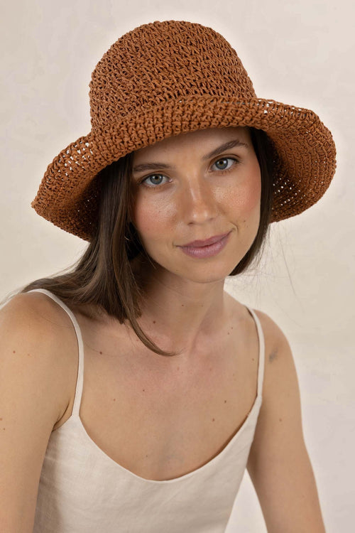 A woman wearing a brown woven sun hat with a wide brim, paired with a light beige spaghetti-strap top, photographed against a neutral beige background.