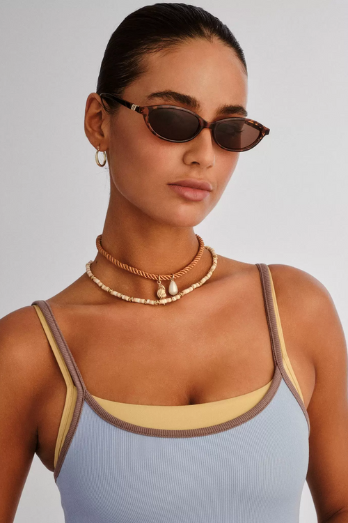Woman wearing sunglasses and a light blue top with brown straps against a gray background