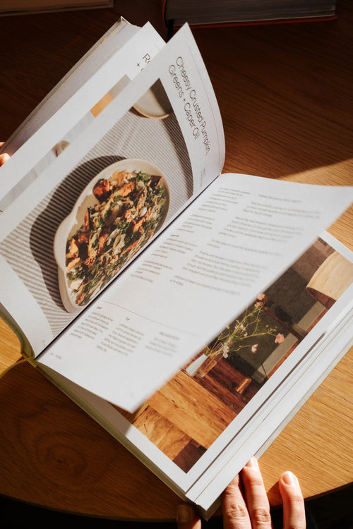 An open cookbook or recipe book spread on a wooden table, with a hand holding the bottom edge. The pages show printed text and food photography, including a dish of pasta.