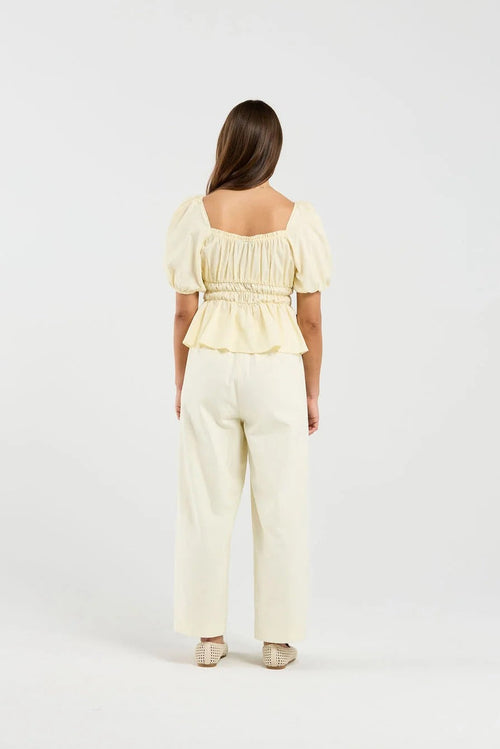 A woman viewed from the back wearing a pale yellow puff-sleeve top with a smocked back and matching cream wide-leg pants, standing against a plain white studio background.