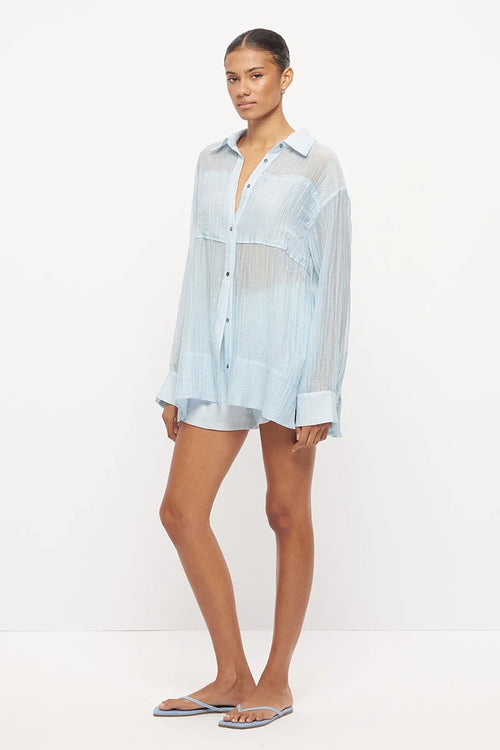 Model wearing a light blue semi-sheer oversized button-up shirt with long sleeves, paired with matching drawstring shorts and flat sandals.