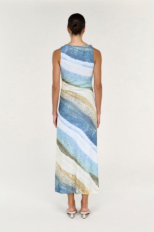 A woman viewed from the back wearing a sleeveless maxi dress with an abstract watercolor-style print in shades of blue, beige, and white, paired with pointed silver heels against a plain light background.