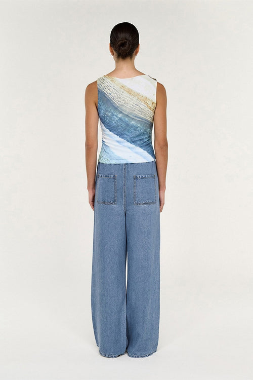 A model shown from the back wearing a sleeveless top with a blue, white, and beige wave print, paired with wide-leg blue denim pants, against a plain light background.