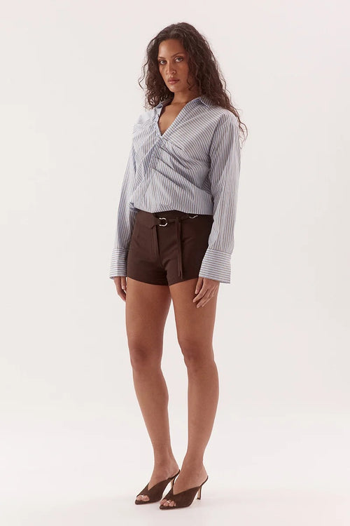A woman modeling a light blue and white striped wrap-style blouse with long sleeves, paired with dark brown shorts and brown mule heels, against a plain white studio background.
