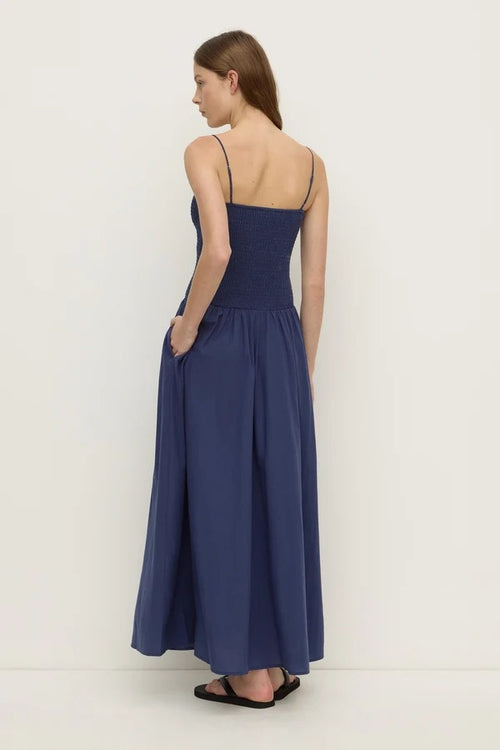 A woman modeling a navy blue maxi dress with thin spaghetti straps, a smocked bodice, and side pockets, shown from a rear three-quarter view against a plain white studio background.