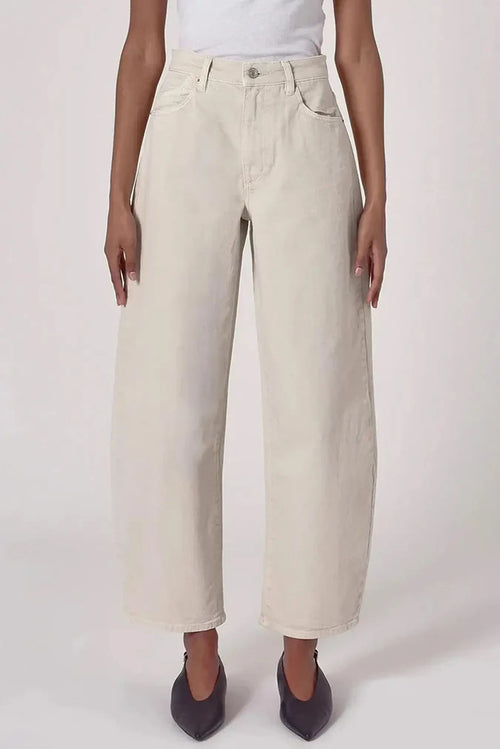 A model wearing a pair of off-white, wide-leg cropped jeans with a white top and dark pointed flats, shown from the waist down against a plain light background.