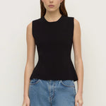 A woman modeling a sleeveless black knit top with a peplum waist, paired with blue jeans, against a plain off-white background.