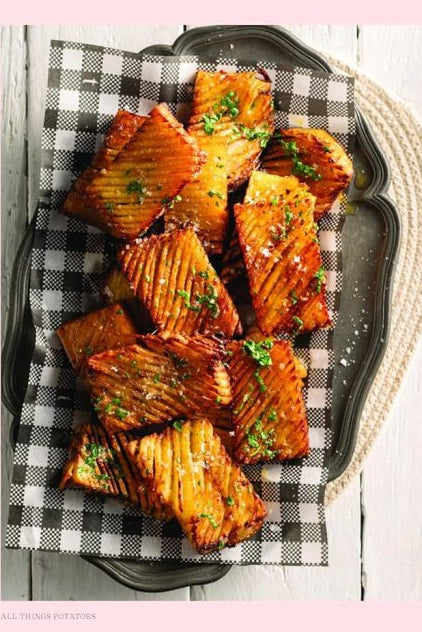 A two-page cookbook spread showing a recipe for Accordion Potatoes. The left page has a pale pink background with the recipe text, and the right page features a photo of glazed potato wraps arranged on a black platter over a black-and-white checkered cloth on a white wooden surface.
