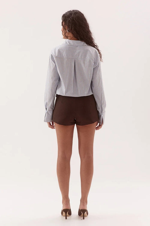A woman viewed from behind wearing a light blue and white vertically striped button-up shirt and dark brown shorts, paired with brown high heels, against a plain white studio background.