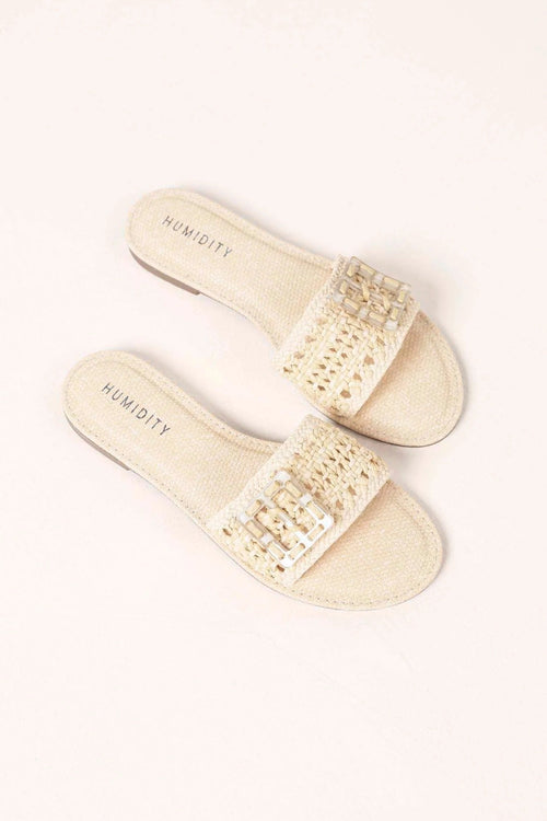 A pair of women's slide sandals in a light cream or off-white woven material, featuring a wide embellished strap with small rhinestone-like studs. The insole is printed with the brand name 'HUMIDITY'.