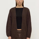 Person wearing a brown cardigan over a black top and brown pants on a white background
