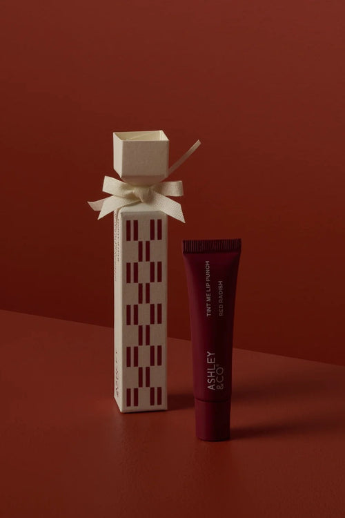 A cosmetic product set featuring a tall cream-colored box with a geometric red pattern and a cream ribbon bow, alongside a matching dark red squeeze tube labeled 'RUE ET ROYAL'. The products are set against a solid dark red background.