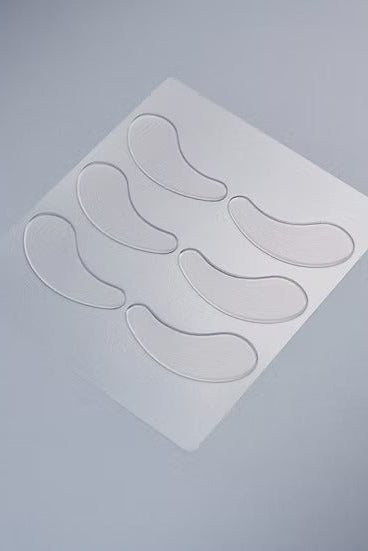 Six clear, teardrop-shaped eye patch pads arranged on a square white backing sheet, photographed on a smooth light gray background.