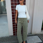 A woman stands outdoors wearing a white ribbed short-sleeve top, olive green wide-leg pants, black flats, and a white fuzzy shoulder bag, posing in front of a building with red-brown tiled columns.