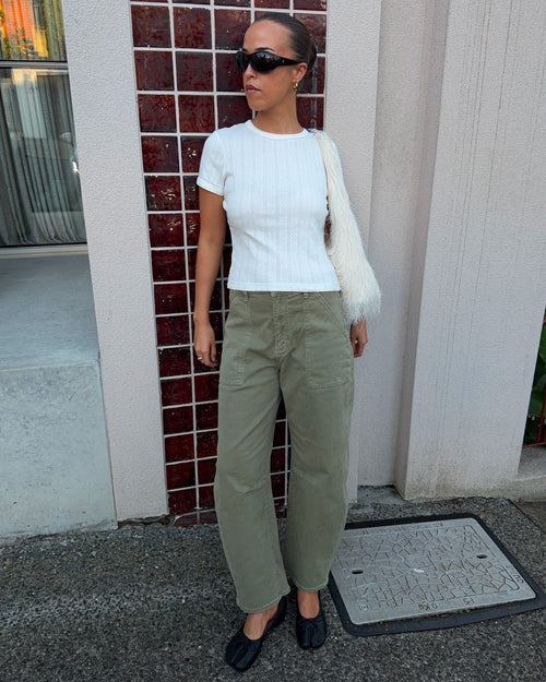 A woman stands outdoors wearing a white ribbed short-sleeve top, olive green wide-leg pants, black flats, and a white fuzzy shoulder bag, posing in front of a building with red-brown tiled columns.