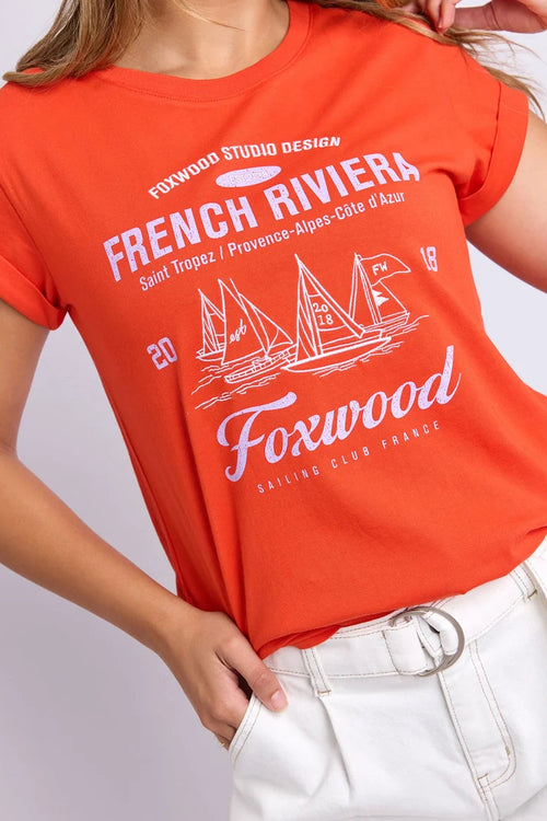 A woman wearing a bright orange short-sleeve graphic T-shirt with white printed text reading 'French Riviera' and sailboat artwork, paired with white pants against a light gray studio background.