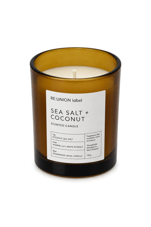An amber glass scented candle in a cylindrical jar with a white label reading 'RE-UNION label SEA SALT + COCONUT SCENTED CANDLE'.