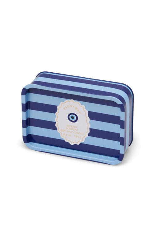 A rectangular tin of Incense + Smoke Eye Blue Stripe 184g Tin Candle, featuring a blue and navy striped pattern with a central cream-colored circular label containing an evil eye design.