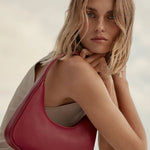 A woman in a beige sleeveless dress carries a large, crescent-shaped burgundy leather shoulder bag with a pebbled texture. The brand name 'MARC JUSTIN' is subtly printed in gold near the bottom right corner of the bag.