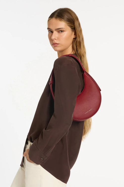 A woman wearing a dark brown button-up shirt and white pants, shown from a side-back angle with a red crescent-shaped shoulder bag slung over her shoulder, against a plain white studio background.
