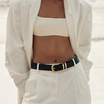 A woman standing on a beach wearing a white blazer, white cropped tube top, white trousers, and a black belt with a gold buckle.