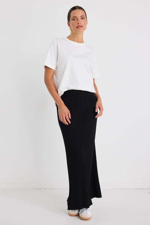 Woman wearing a white boxy tee tucked into a full-length black rib knit skirt with flared hem, styled with white sneakers.