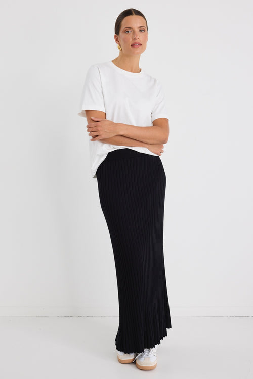 Woman wearing a white boxy tee tucked into a full-length black rib knit skirt with flared hem, styled with white sneakers.