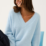 A woman wearing a fuzzy light blue V-neck sweater and dark patterned pants, seated on a light blue chair against a plain white background.