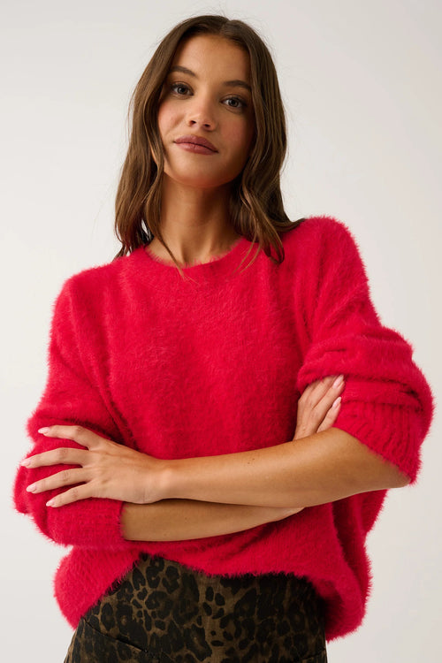 A woman wearing a fuzzy bright pink crewneck sweater and leopard-print pants, posing with her arms crossed against a plain light gray background.